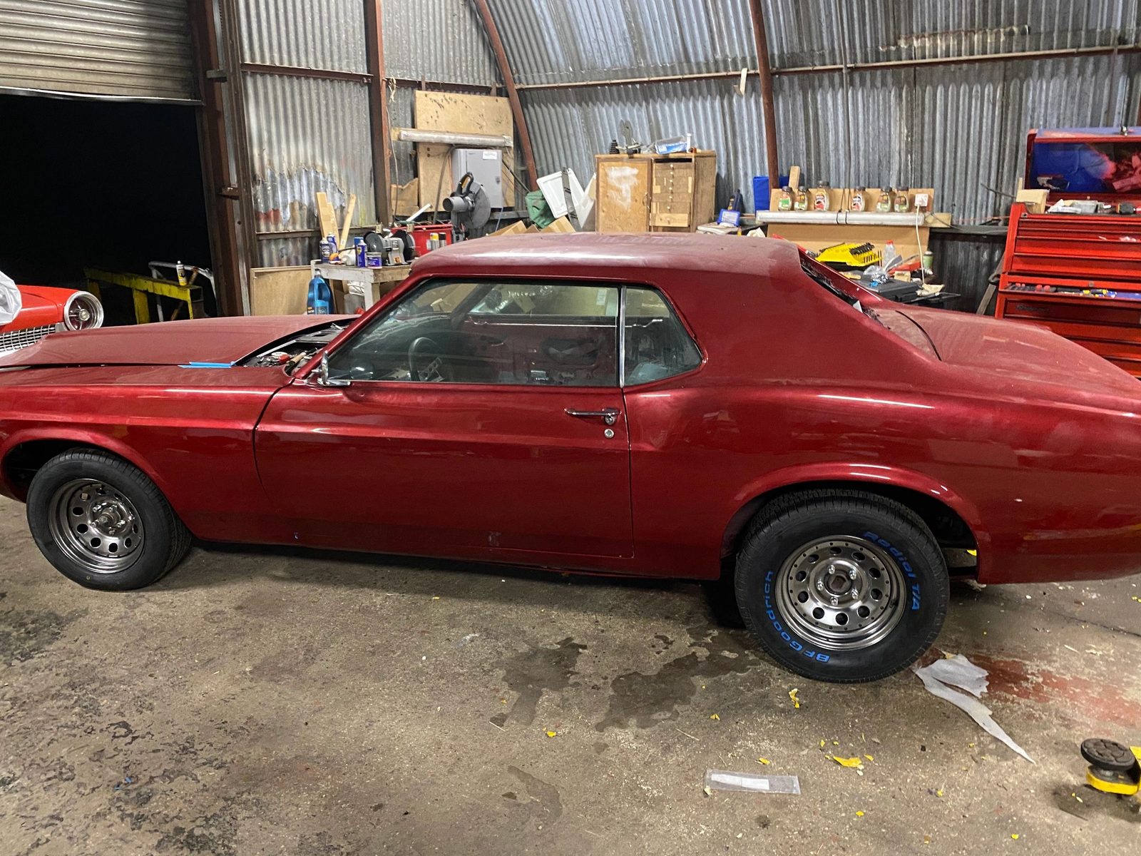 Red Mustang coupe side-on inside the workshop, with tools and storage behind it
