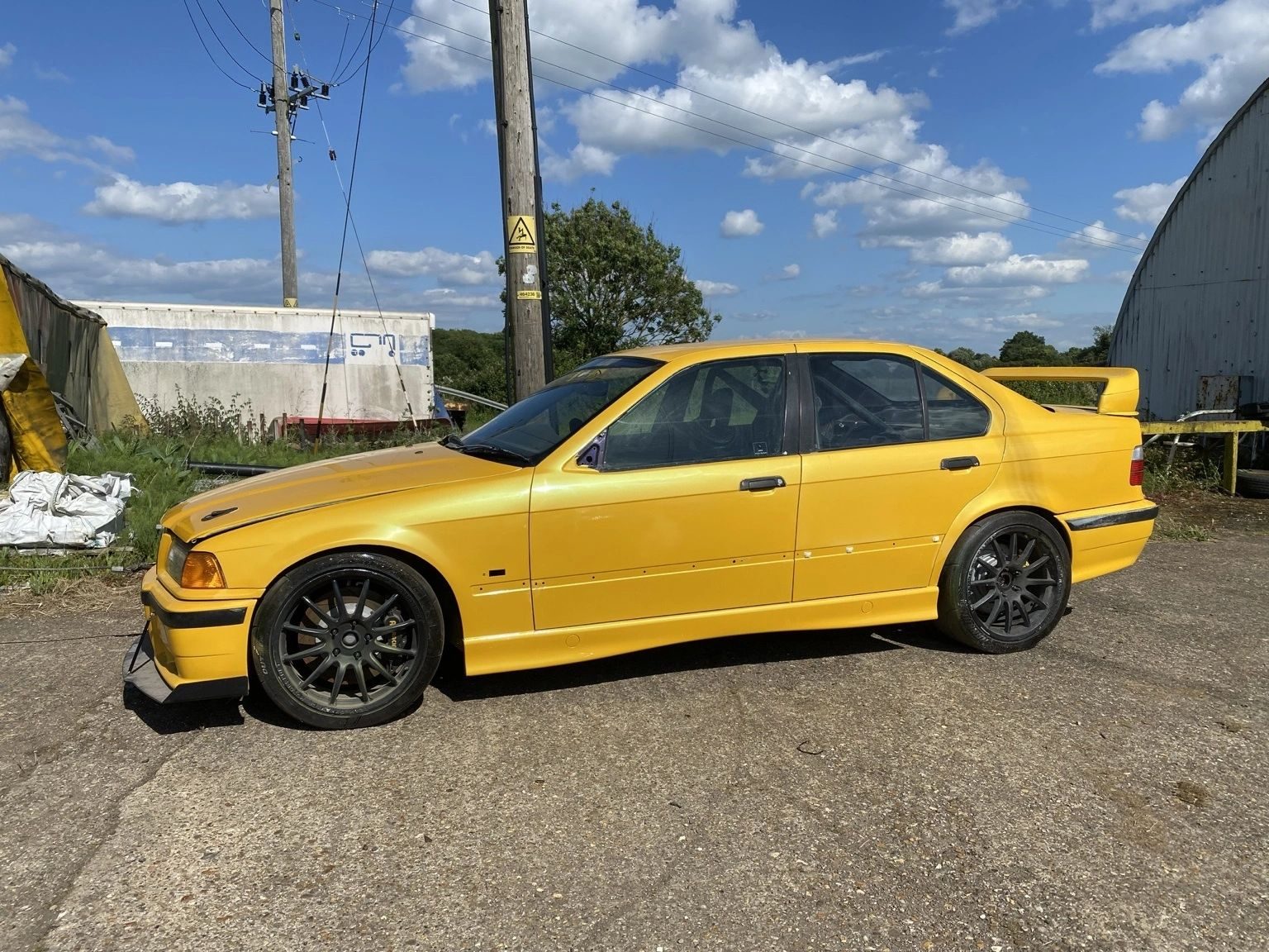 Yellow BMW E36 M3 saloon with black alloy wheels and a front splitter, parked outdoors